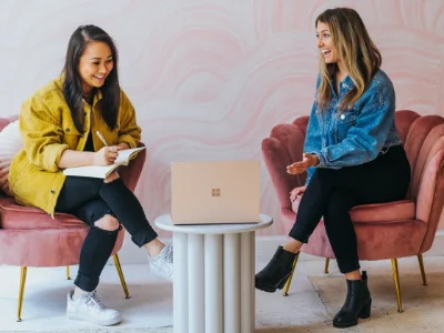 Zwei Frauen sitzen nebeneinander auf zwei Stühlen und schauen in einen Laptop der in der Mitte des Raumes steht