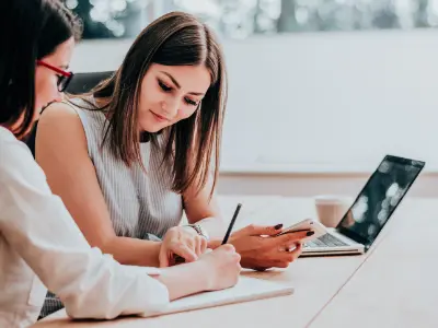 Zwei Frauen diskutieren an einem Tisch mit Laptop und Notebook über Gesundheitsmanagement.