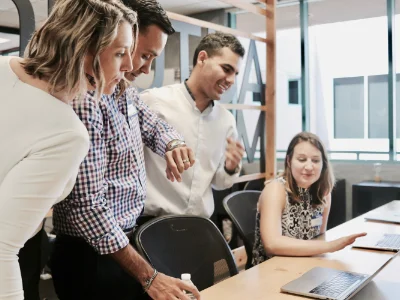 Vier internationale Manager:innen sind in einem Besprechungsraum und schauen in zwei Laptops. Dabei schaut ein Mann in Anzug auf die Uhr
